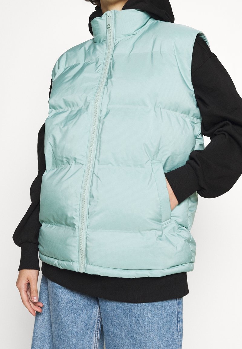 Personne portant un gilet matelassé bleu clair par-dessus un sweat à capuche noir et un jean bleu, main dans la poche du gilet, sur fond blanc uni.