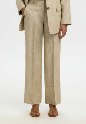 Beige bukser med vide ben og tynde striber, kombineret med matchende blazer og brune åbne sandaler på en stående person.
