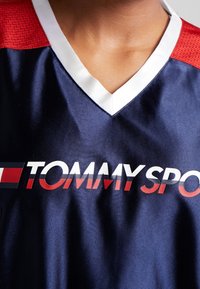 Camiseta deportiva Tommy Sport azul marino con detalles en malla roja en los hombros, cuello en V blanco y un logo impreso en negrita en el pecho. Textura suave.