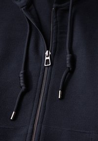 Veste à capuche zippée marine avec un matériau texturé, une fermeture éclair argentée et des cordons avec des embouts en métal. Design classique avec une coupe ajustée.