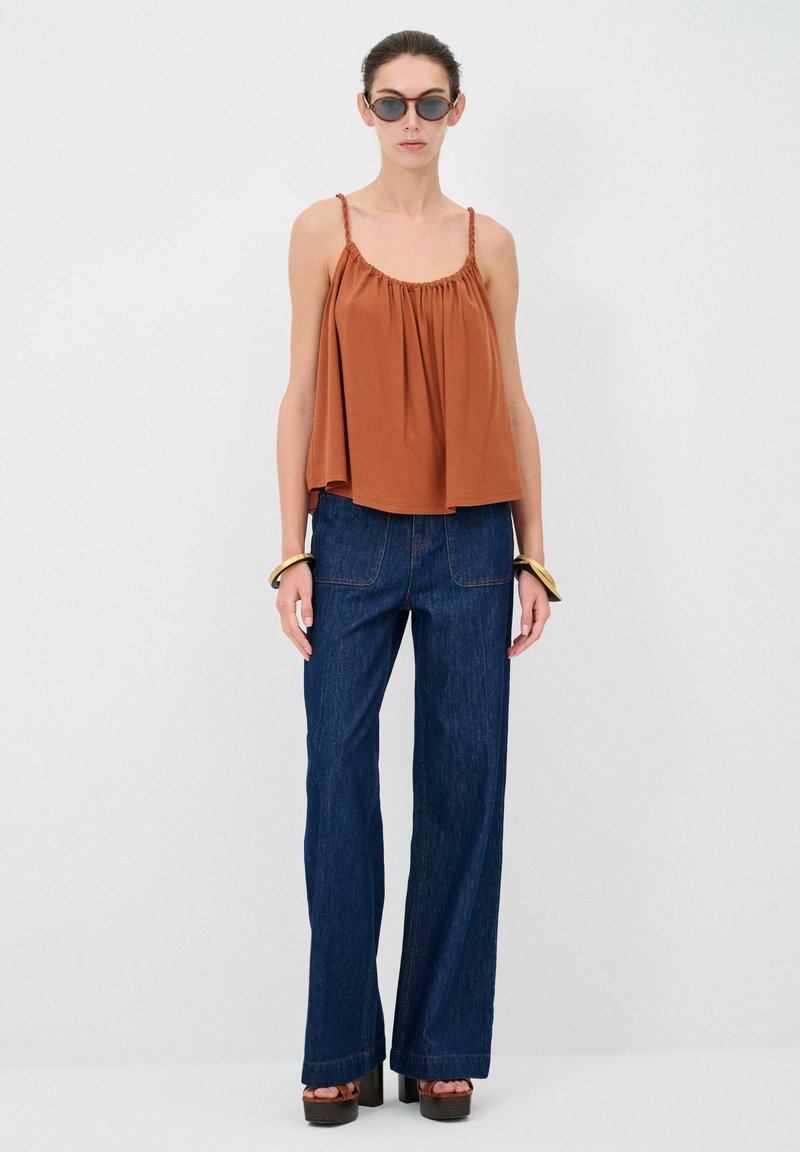 Top fluido marrone con spalline regolabili; abbinato a jeans a vita alta, larghi e blu scuro; modello che indossa occhiali da sole tondi e bracciali spessi.