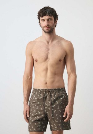 Man met kort krullend donker haar, die bruine zwemshorts met een licht paisleypatroon draagt, staand tegen een effen achtergrond.