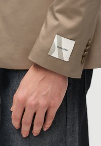 Χέρι ακουμπισμένο στο πλάι, φορώντας σακάκι καμηλό με ετικέτα Calvin Klein στον μανίκι και σκούρο τζιν.