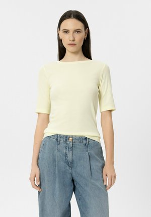 Frau mit glattem dunklem Haar, die ein blassgelbes Oberteil mit Ellbogenärmeln und hoch taillierte, lockere blaue Jeans trägt, steht vor einem weißen Hintergrund.