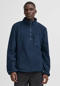 Maglione in pile navy con colletto alto, zip a quarto, tasca frontale e tasche laterali. Caratterizzato da una consistenza morbida e una vestibilità rilassata.