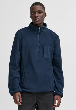 Marineblå fleece genser med høy krage, kvart glidning, frontlomme og sidelommer. Har en myk tekstur og en avslappet passform.