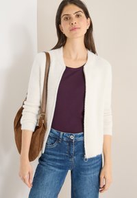 Biały miękki kardigan z okrągłym dekoltem, noszony na ciemnofioletowej bluzce; zestawiony z niebieskimi dżinsami i brązową zamszową torebką na ramię.