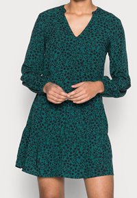 Grön klänning med V-ringning, långa ärmar och flerlagad kjol. Har ett blommönster i mörka nyanser och är tillverkad av ett lätt tyg.