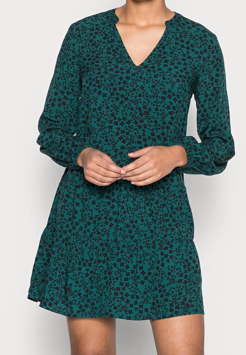 Grön klänning med V-ringning, långa ärmar och flerlagad kjol. Har ett blommönster i mörka nyanser och är tillverkad av ett lätt tyg.