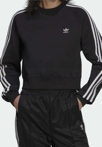 Svart sweatshirt med vita ränder på ärmarna, rundad halsringning och logodetalj. Matchas med svarta byxor i ett elegant, slätt material.