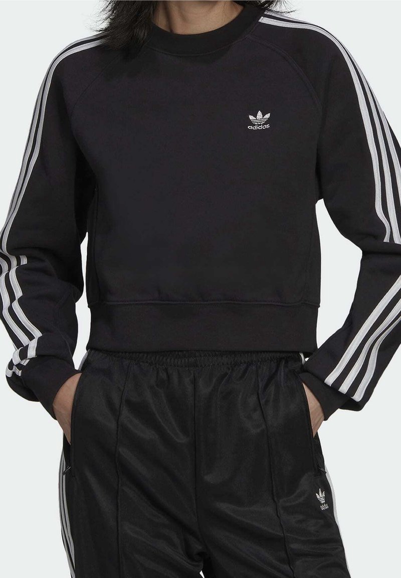 Svart sweatshirt med vita ränder på ärmarna, rundad halsringning och logodetalj. Matchas med svarta byxor i ett elegant, slätt material.