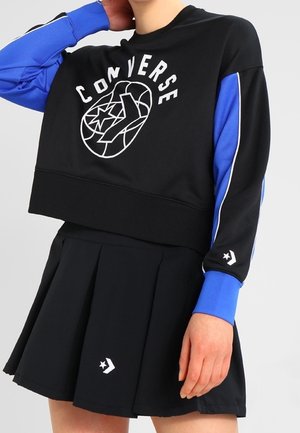 Kobieta ubrana w czarną krótką bluzę Converse z niebieskimi rękawami oraz dopasowaną czarną plisowaną spódnicę z logo Converse w kształcie gwiazdy chevron.