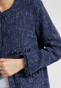 Tummansininen tweed-takki, jossa on teksturoitu kuosi, frayed-hihansuut ja yksinkertainen kaula-aukko. Ei näkyviä kiinnityksiä tai koristeluja.