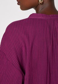 Tessuto a righe viola con una texture a costine, caratterizzato da un collo a barca e una silhouette ampia, accentuato da una cucitura piegata sul retro.