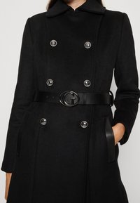 Vrouw in een zwarte dubbelzijdige jas met zilveren knopen en een zwarte riem met een "G" gesp, met één hand in een zak.