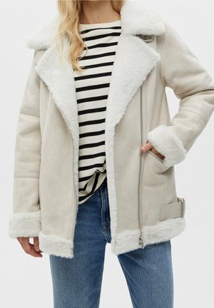 Kvinde iført en beige fåreskindsjakke med hvid pelskant, sort-og-hvid stribet skjorte og blå jeans, hånd i jakkelommen.
