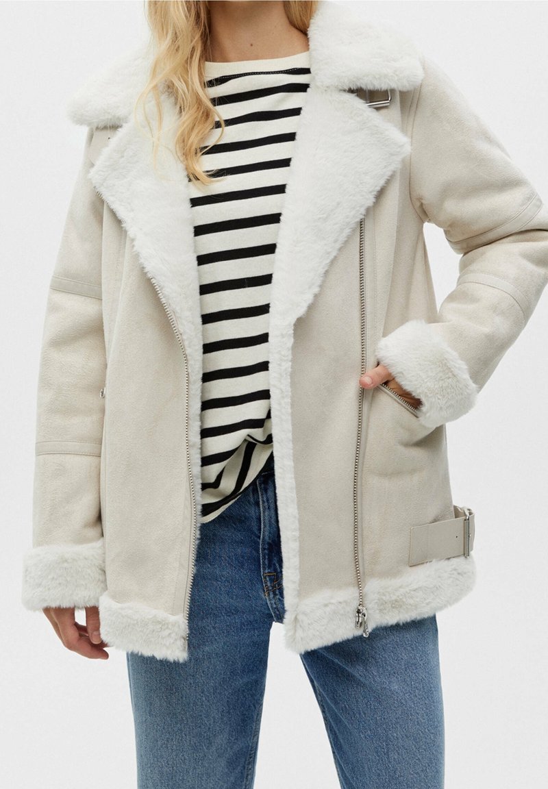 Femme portant une veste en peau retournée beige avec une bordure en fourrure blanche, une chemise rayée noir et blanc, et un jean bleu, main dans la poche de la veste.