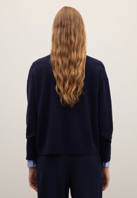 Marineblauer Pullover mit lockerem Schnitt, gerippten Bündchen und Saum. Lange, wellige Haare sind sichtbar, kombiniert mit hellblauen Hemdmanschetten.