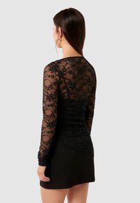 Μαύρη δαντελένια μπλούζα με μακριά μανίκια και διαφανές ύφασμα με λουλουδάτα σχέδια, επενδυμένη πάνω σε μια στενή μπλούζα σε σταθερό μαύρο χρώμα, συνδυασμένη με μια μαύρη φούστα.