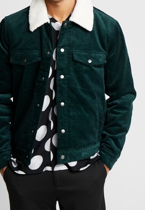 Man draagt een donkergroene corduroy jas met witte fleece kraag over een zwart-wit gestippeld overhemd en zwarte broek, handen in de zakken.