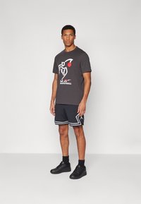 Grå t-shirt med en basketballgrafik og røde detaljer, parret med sorte shorts med hvide striber og sorte sneakers.