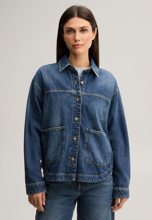 Femme aux cheveux bruns raides portant une veste en jean bleue avec des boutons et de grandes poches avant, associée à un jean bleu.