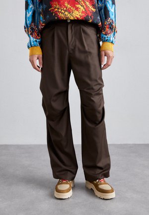 Brown, wide-leg trousers made from lightweight fabric with front pleats. Worn with chunky beige and brown sneakers featuring red accents.