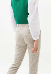 Pantalon à carreaux beige clair avec une ceinture plate et deux poches arrière, associé à un gilet en tricot vert porté sur une chemise blanche à manches longues.
