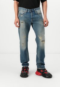 Jeans in denim azzurro chiaro con taglio dritto, caratterizzati da toppe strappate e scoloriture. Indossati con sneakers sportive nere e rosse.