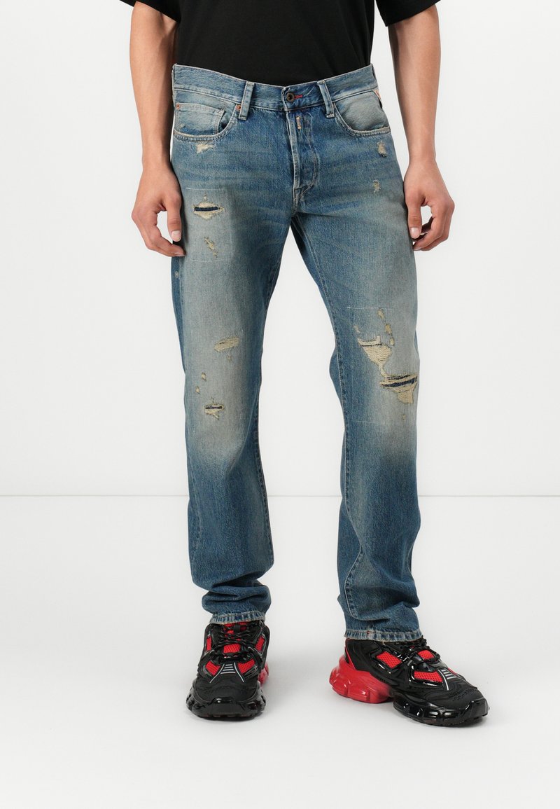 Lyseblå denim jeans med lige snit, prydet med slidte lapper og falmede områder. Båret med sorte og røde træningssko.