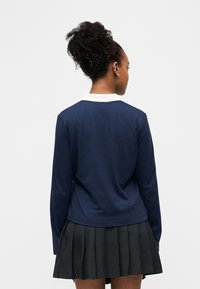 Femme aux cheveux bouclés attachés en chignon, portant un haut bleu marine à manches longues avec un col blanc et une jupe plissée foncée, vue de dos.