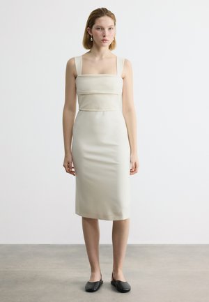 Frau steht und trägt ein ärmelloses, knielanges, figurbetontes cremefarbenes Kleid mit quadratischem Ausschnitt und schwarze Slip-On-Schuhe vor einem schlichten Hintergrund.