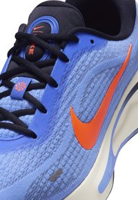Tenisky Nike s modrým texturovaným svrškem, oranžovým logem swoosh, černými tkaničkami a polstrovaným límcem. Mezi pozoruhodné detaily patří kulaté logo a lehká podrážka.