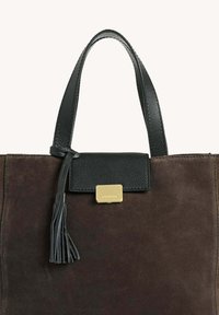Sac fourre-tout en daim marron foncé avec une anse en cuir noir, un rabat, un fermoir rectangulaire en or et un détail de pompon en cuir noir.