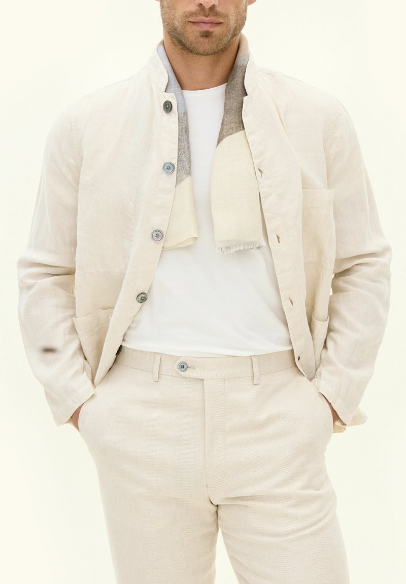 Beige linnen jas met knoopsluiting, staande kraag en zijzakken, gedragen over een wit shirt en bijpassende beige broek.