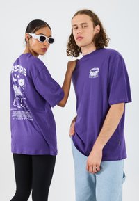 Camisetas oversized moradas con estampados gráficos en el frente y en la parte de atrás. Modelo usando gafas de sol blancas; un modelo en leggings negros, otro en denim claro.