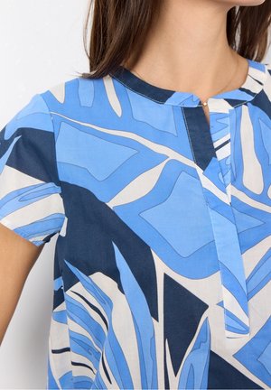 Nahaufnahme einer Frau, die eine blaue, gemusterte Kurzarmbluse mit abstrakten Blattmustern und einem geteilten Halsausschnitt trägt.