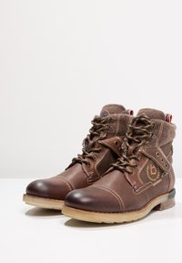 Botas de cuero marrón hasta el tobillo con collar de forro polar, puntera redonda, costuras contrastantes y suela de goma. Ojetes de metal para los cordones y detalle del logo.