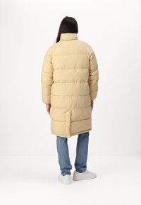 Beige pufferjack met een hoge kraag, horizontale stiksels en zijsplitten, gedragen over blauwe jeans en witte sneakers. Lijkt warm en casual.