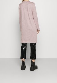 Lång, ljusrosa tröja med rundad halsringning, i kombination med glänsande svarta capribyxor och kraftiga svarta ankelstövlar. Slät textur totalt sett.