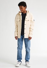 Chaqueta crema con capucha y botones en el frente, con dos bolsillos en el pecho, usada sobre una camisa negra, acompañada de jeans azules y zapatillas blancas.