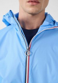 Hellblaue Jacke mit durchgehendem Reißverschluss, kontrastierendem rotem und blauem Reißverschlussband, verstellbarer Kapuze und glatter, glänzender Textur.