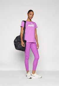 Rožinė PUMA marškinėlių su balta logo, derinami su raštuotomis purpurinėmis leggins. Juoda sportinė kuprinė su petnešomis, balti ir rusvi sportbačiai.