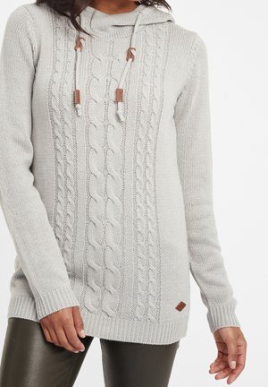 Femme portant un pull à capuche gris clair en maille torsadée avec cordons et pantalon foncé, debout devant un fond blanc.