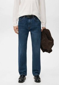 Vaqueros de mezclilla azul oscuro con corte recto, diseño de cinco bolsillos y costuras visibles. Combinados con una camisa blanca de manga larga y calzado negro.