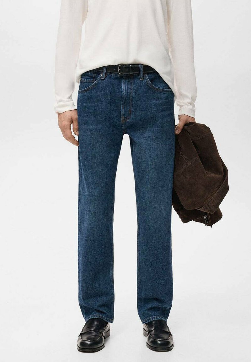 Vaqueros de mezclilla azul oscuro con corte recto, diseño de cinco bolsillos y costuras visibles. Combinados con una camisa blanca de manga larga y calzado negro.