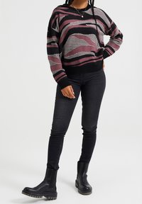 Un suéter a rayas colorido en tonos de negro, rosa y gris, con un corte holgado y puños y dobladillo de canalé, combinado con jeans oscuros y botas negras gruesas.