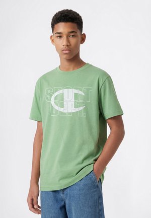 Teenage dreng med kort krøllet hår iført en grøn t-shirt med teksten "SPORT DEPT." og blå jeans står mod en hvid baggrund.