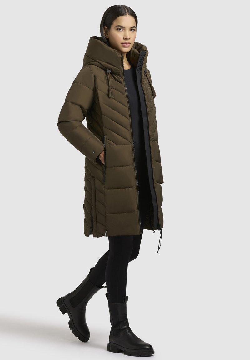 Padded long coat in oliwkowym kolorze z kapturem, z chevronowym przeszyciem, kieszeniami na zamek oraz luźnym krojem. Noszony z czarnymi legginsami i botkami.
