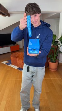 Blaues MoMA-Box mit glatter Oberfläche, rechteckiger Form und einer runden Öffnung oben. Verfügt über ein passendes blaues Schlüsselband.